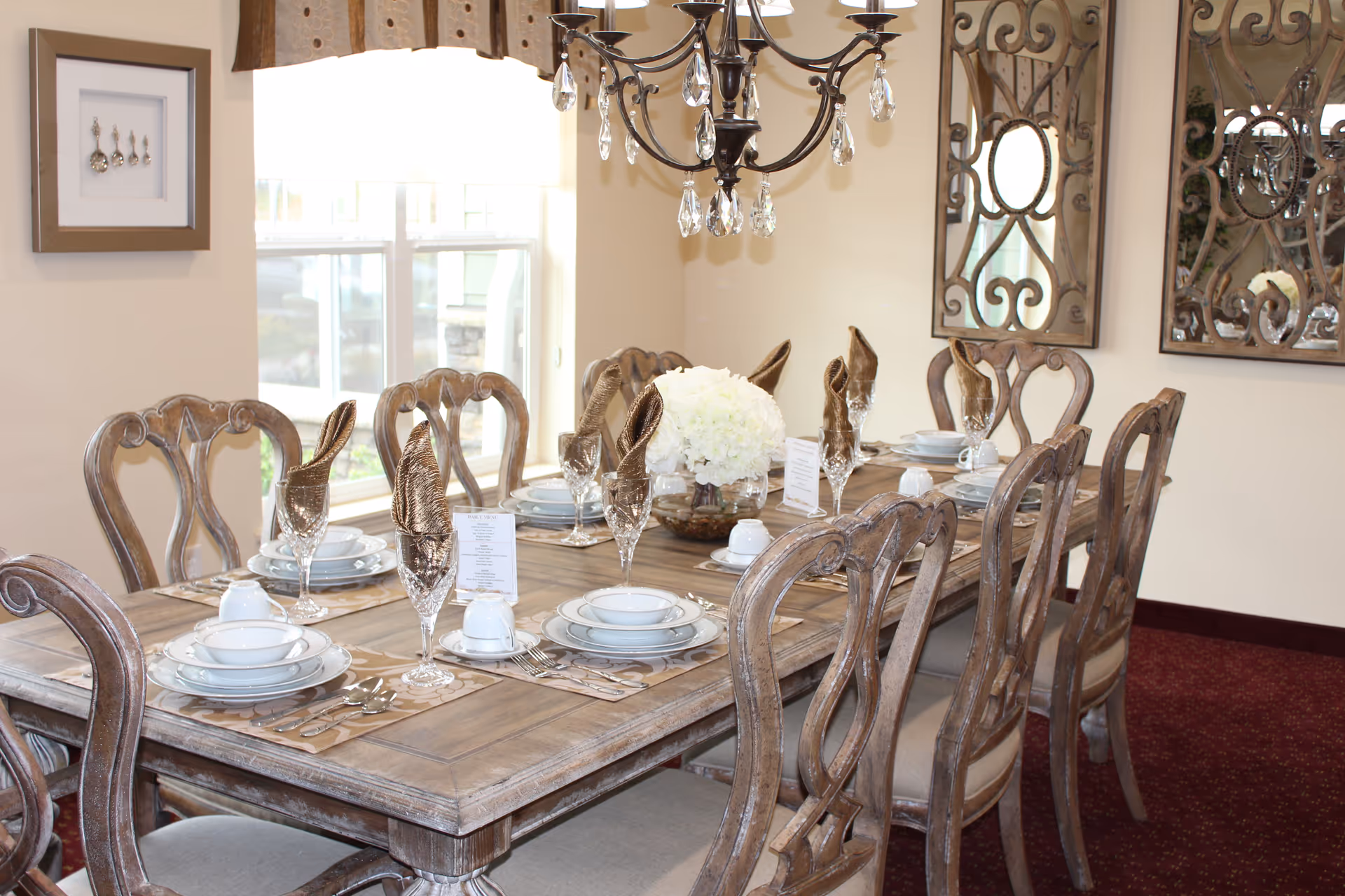 A formal dining room with a long wooden table set with plates, glasses, napkins, and a floral centerpiece beneath a chandelier.