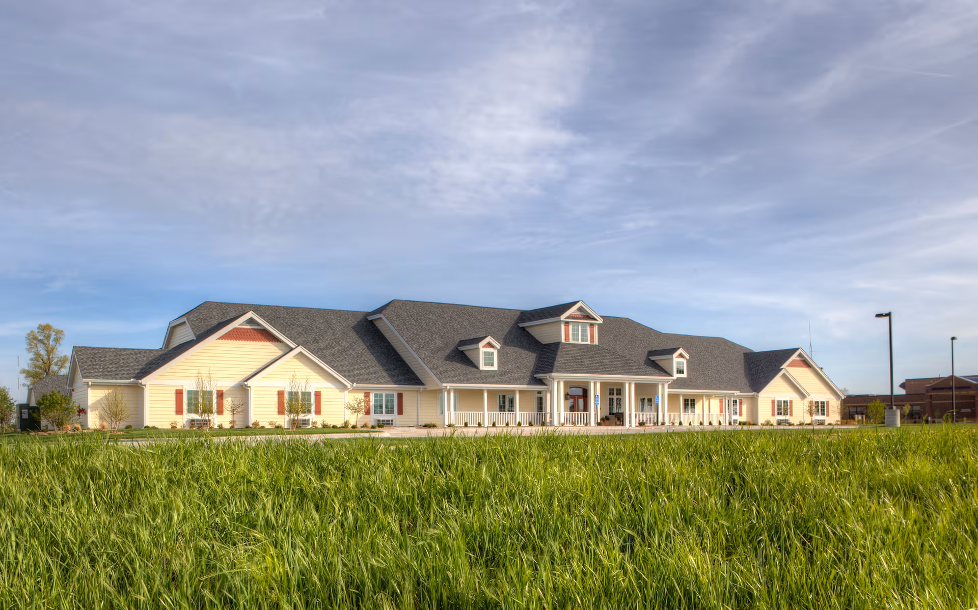 Wide exterior view of a single-story assisted living facility building with a gray roof and light yellow siding, surrounded by green grass under a partly cloudy sky.