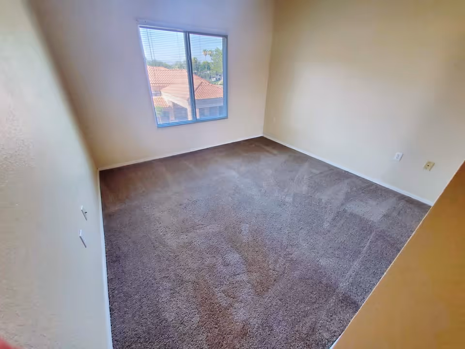 Empty room with beige walls and brown carpet flooring, a window with blinds showing a view of rooftops and trees outside.