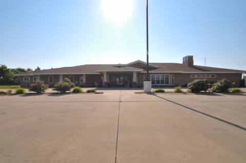 Front exterior view of a single-story building with a wide driveway and landscaped bushes in front under a clear blue sky.