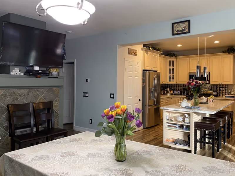 Interior view of a home showing a kitchen and dining area. The foreground features a table with a floral tablecloth and a vase of colorful tulips. The kitchen has cream-colored cabinets, a stainless steel refrigerator, and an island with bar stools. A TV is mounted above a fireplace with two wooden chairs in front of it. The walls are painted light blue, and there are decorative items on the kitchen island and above the cabinets.