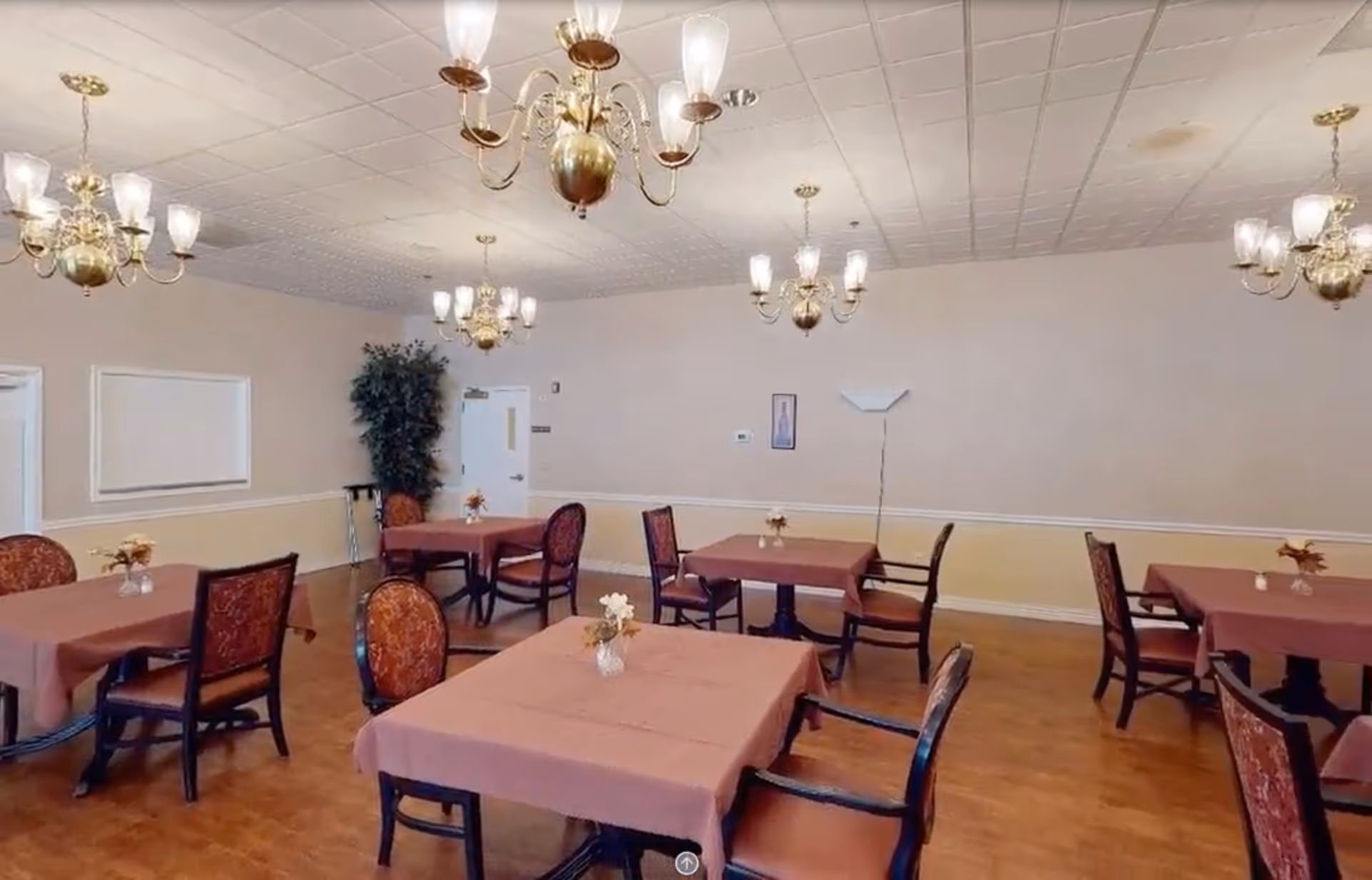 A dining room with several square tables covered with pink tablecloths, each table surrounded by chairs with patterned upholstery. The room has wooden flooring, beige walls with white trim, multiple brass chandeliers with glass shades hanging from a white tiled ceiling, a tall potted plant in the corner, and a floor lamp against the wall.