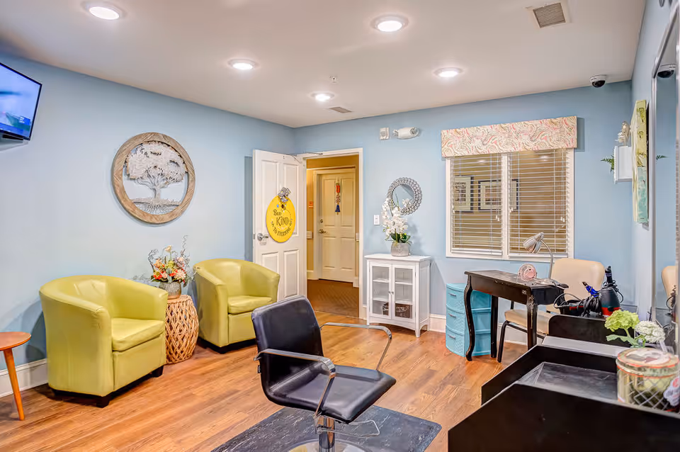 Bright communal salon/waiting area with green armchairs, a black salon chair, small tables and a desk against blue walls.