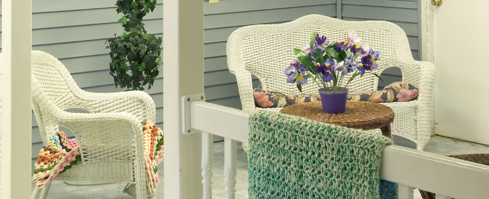 A cozy outdoor seating area with white wicker chairs and a matching loveseat, adorned with floral cushions and colorful knitted blankets. A small round wicker table holds a purple pot with vibrant purple and yellow flowers. The area is enclosed by light gray siding walls and white railings.