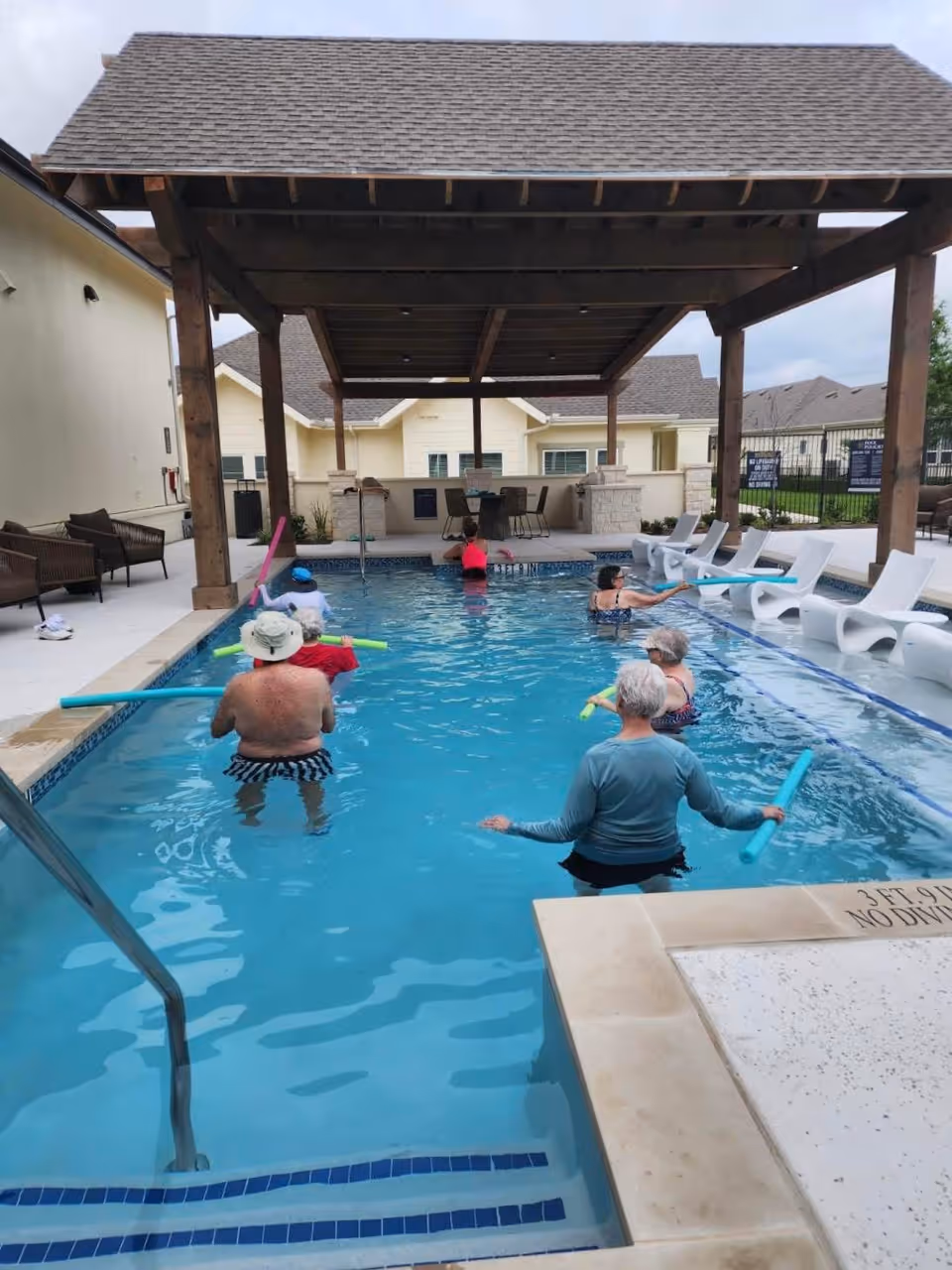 A group of elderly people participating in a water exercise class in an outdoor swimming pool under a wooden pergola. The pool area has white lounge chairs on the right side and some seating on the left side. Residential buildings are visible in the background.