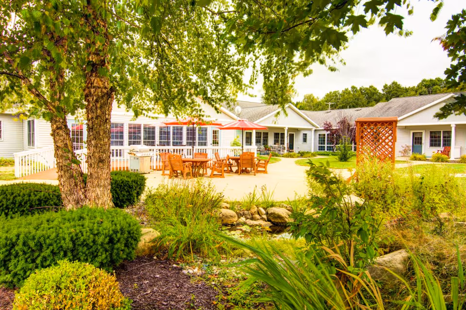 Outdoor patio area at Curry House featuring wooden tables and chairs with red umbrellas, surrounded by greenery, bushes, trees, and a small pond in the foreground. The building with white siding and multiple windows is visible in the background under a cloudy sky.