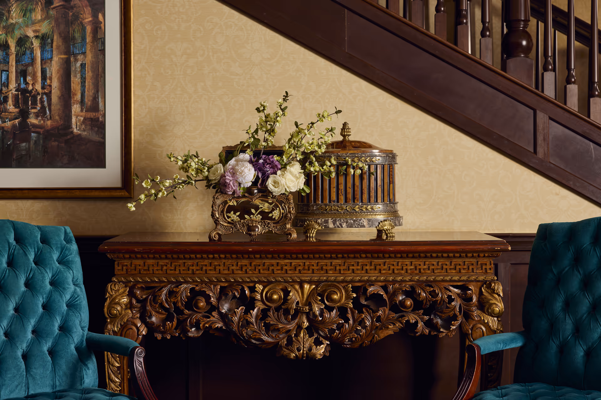 An ornately carved wooden console table with gold accents is positioned against a patterned beige wall beneath a dark wooden staircase. On the table, there is a decorative floral arrangement with white and purple flowers in an ornate vase, and a vintage-style birdcage. Two teal tufted armchairs flank the table. A framed painting depicting a scene with columns and people is partially visible on the left side of the image.