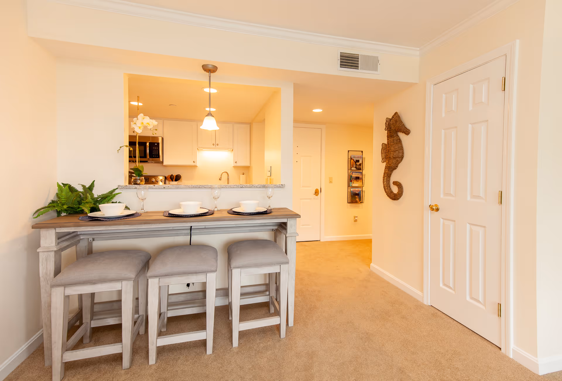 Interior view of a senior living facility showing a small dining area with a wooden table and three cushioned stools. The table is set with placemats, bowls, and wine glasses. Behind the table is a kitchen with white cabinets, a microwave, and a sink. The walls are painted light beige, and there is a decorative wicker seahorse hanging on the wall near a closed white door. The floor is carpeted in a light tan color.