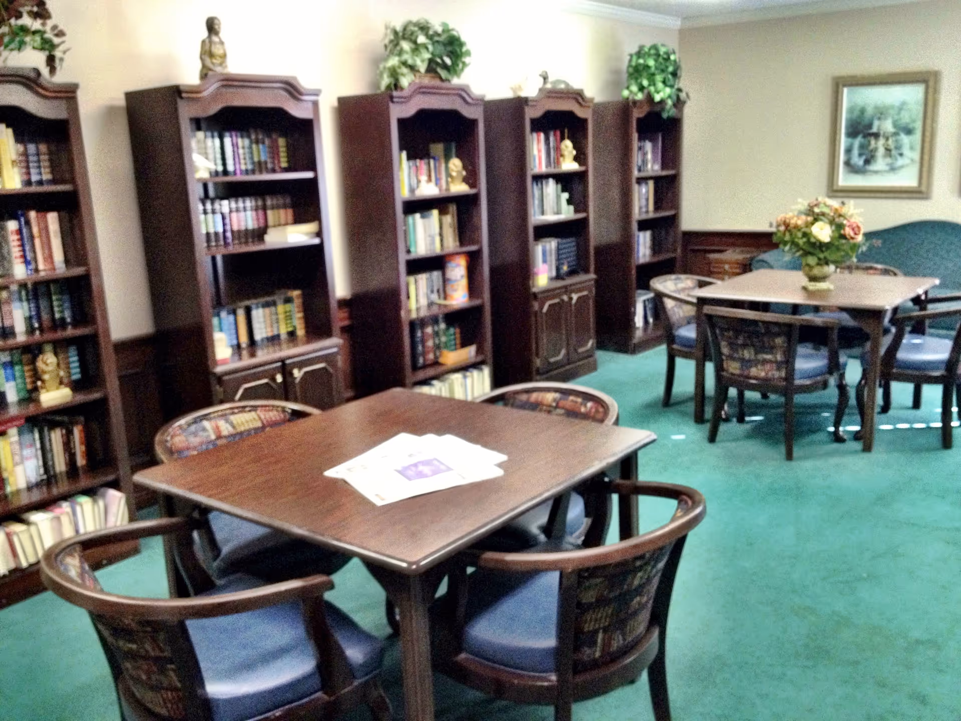 A cozy interior room with green carpet featuring two wooden tables surrounded by cushioned chairs. Along the wall are several tall wooden bookshelves filled with books and decorative items. A framed picture hangs on the wall above a green upholstered bench, and a vase with flowers is placed on one of the tables.