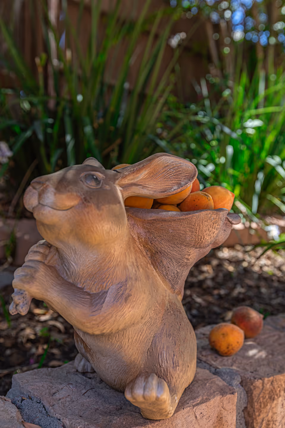 A decorative statue of a rabbit holding a basket filled with peaches, placed outdoors on a stone surface with green plants and a wooden fence in the background.