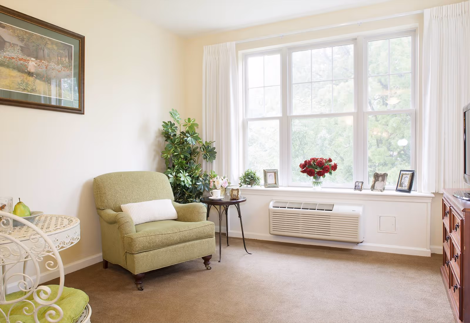 A cozy living room area with a green upholstered armchair and a small round side table holding a teacup and flowers. There is a large window with white curtains letting in natural light, decorated with framed photos and a vase of red flowers on the windowsill. A wooden dresser with a TV on top is visible on the right side, and a framed painting hangs on the wall above the armchair. The room has beige carpet and light-colored walls.