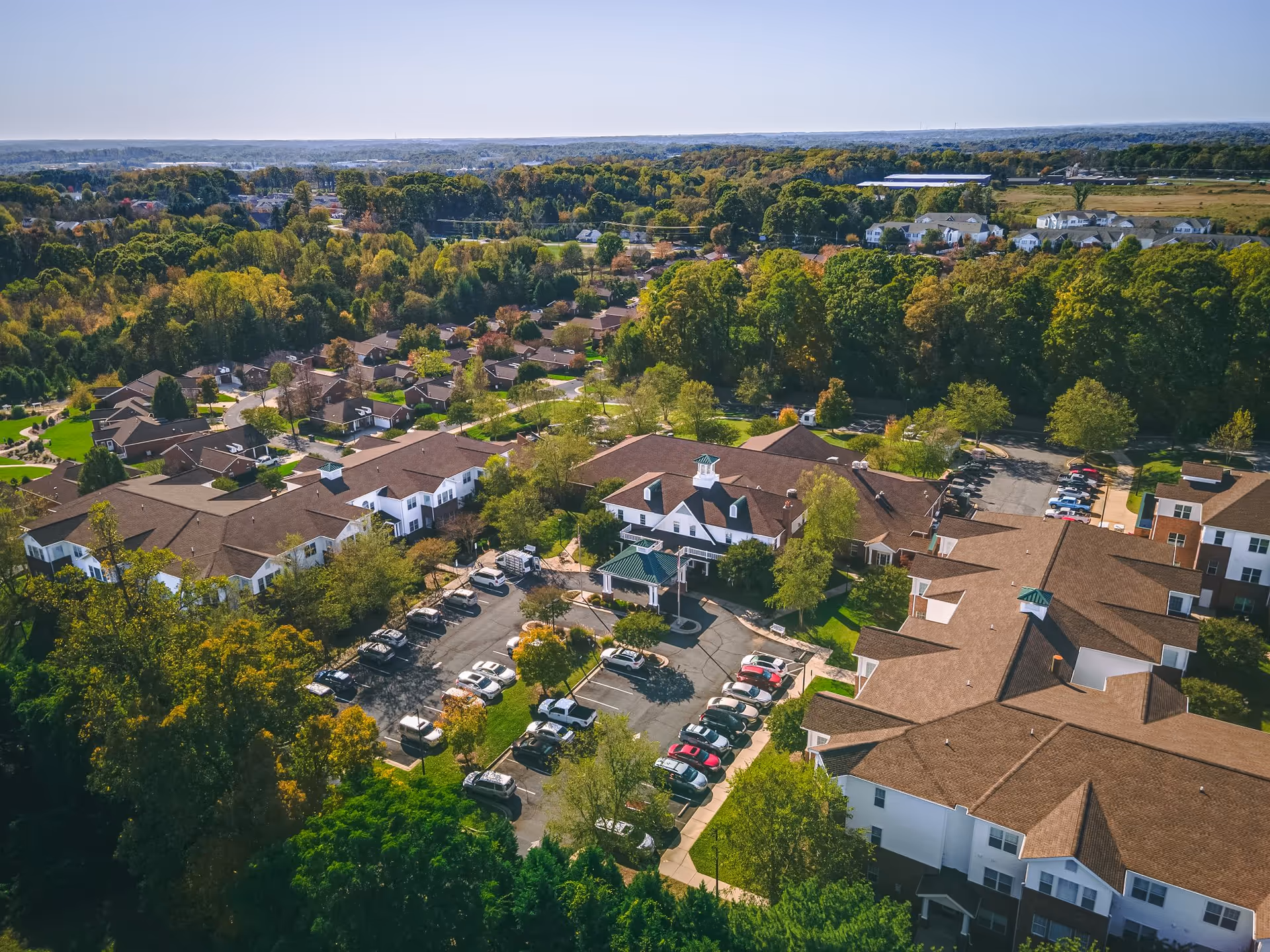 Aerial view of Homestead Hills senior living facility surrounded by trees and greenery. The image shows multiple buildings with brown roofs, parking lots with cars, and a large wooded area in the background under a clear sky.