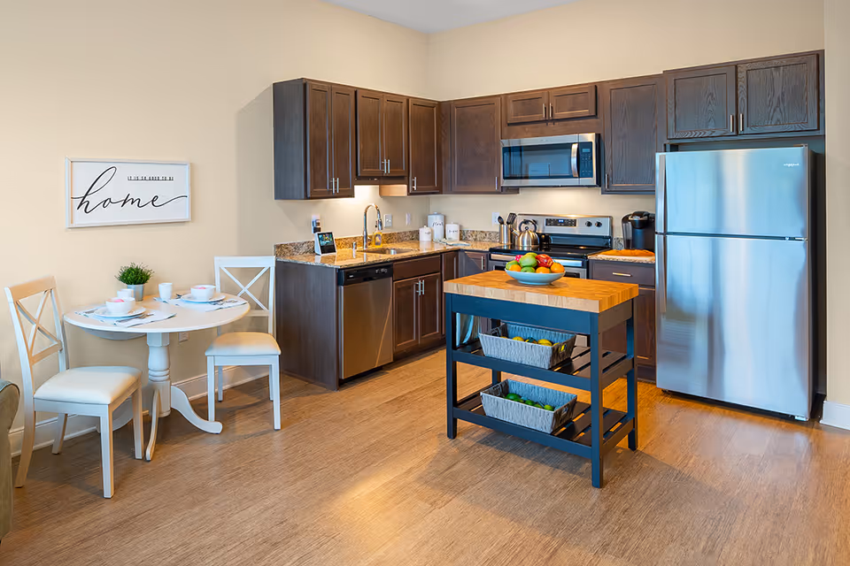 A modern kitchen with dark wood cabinets, stainless steel appliances including a refrigerator, microwave, stove, and dishwasher. There is a small kitchen island with a wooden top and two baskets underneath, holding fruits and vegetables. To the left, there is a small round dining table set for two with white chairs and a small plant on the table. A wall decoration with the word 'home' is hung above the dining area.