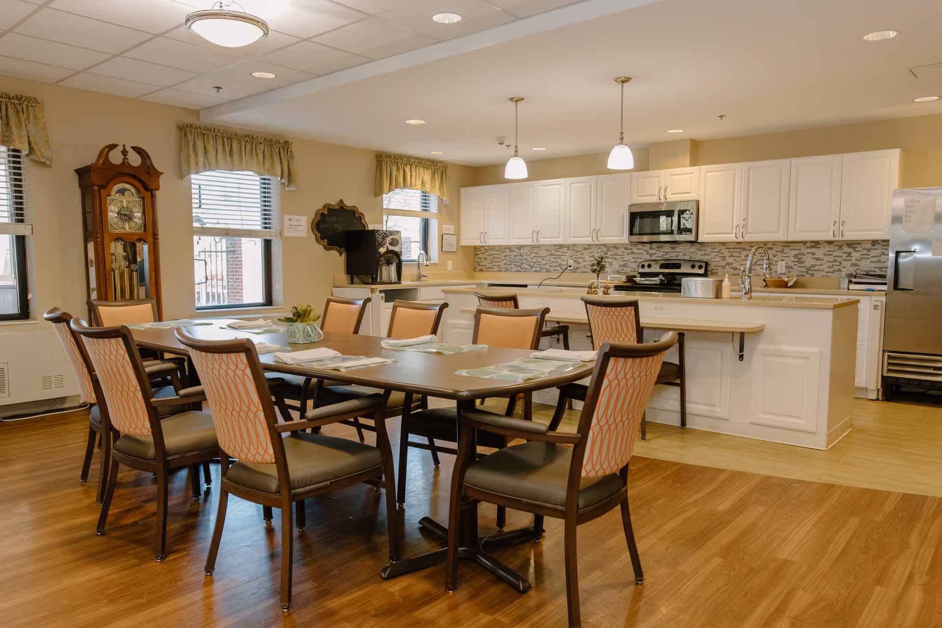 Dining area with a large table and chairs in front of an open kitchen with white cabinets and stainless steel appliances.