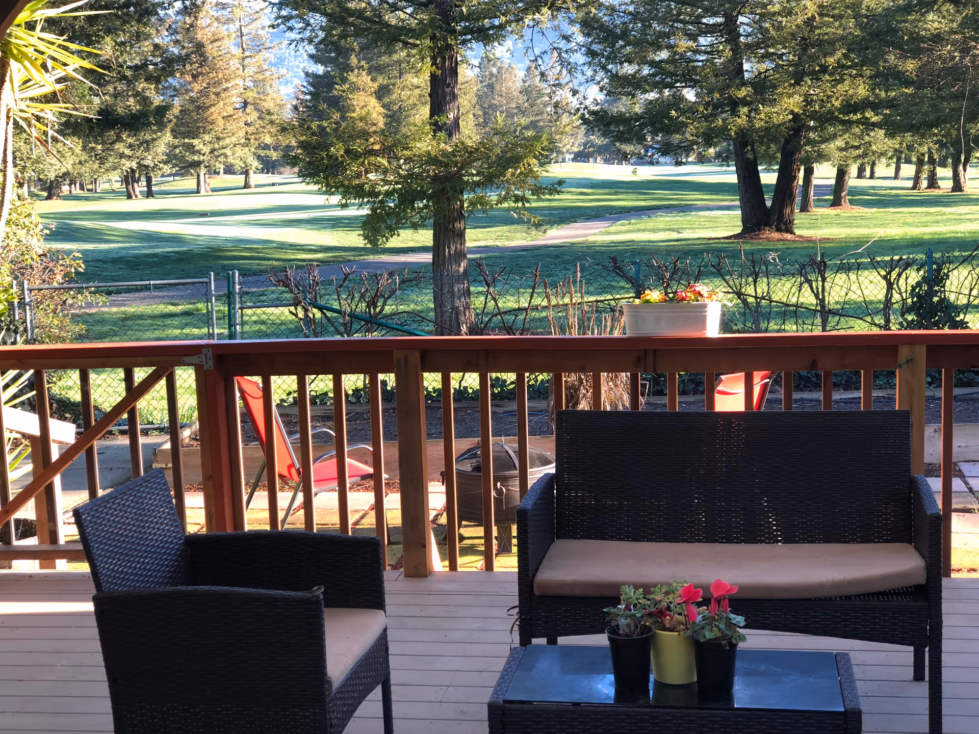 Outdoor patio area with a wooden deck featuring a black wicker loveseat with beige cushions, a matching armchair, and a glass-top coffee table with three small potted plants. Beyond the deck railing, there is a fenced yard with a fire pit and red chairs, and a green grassy area with trees in the background under a clear sky.