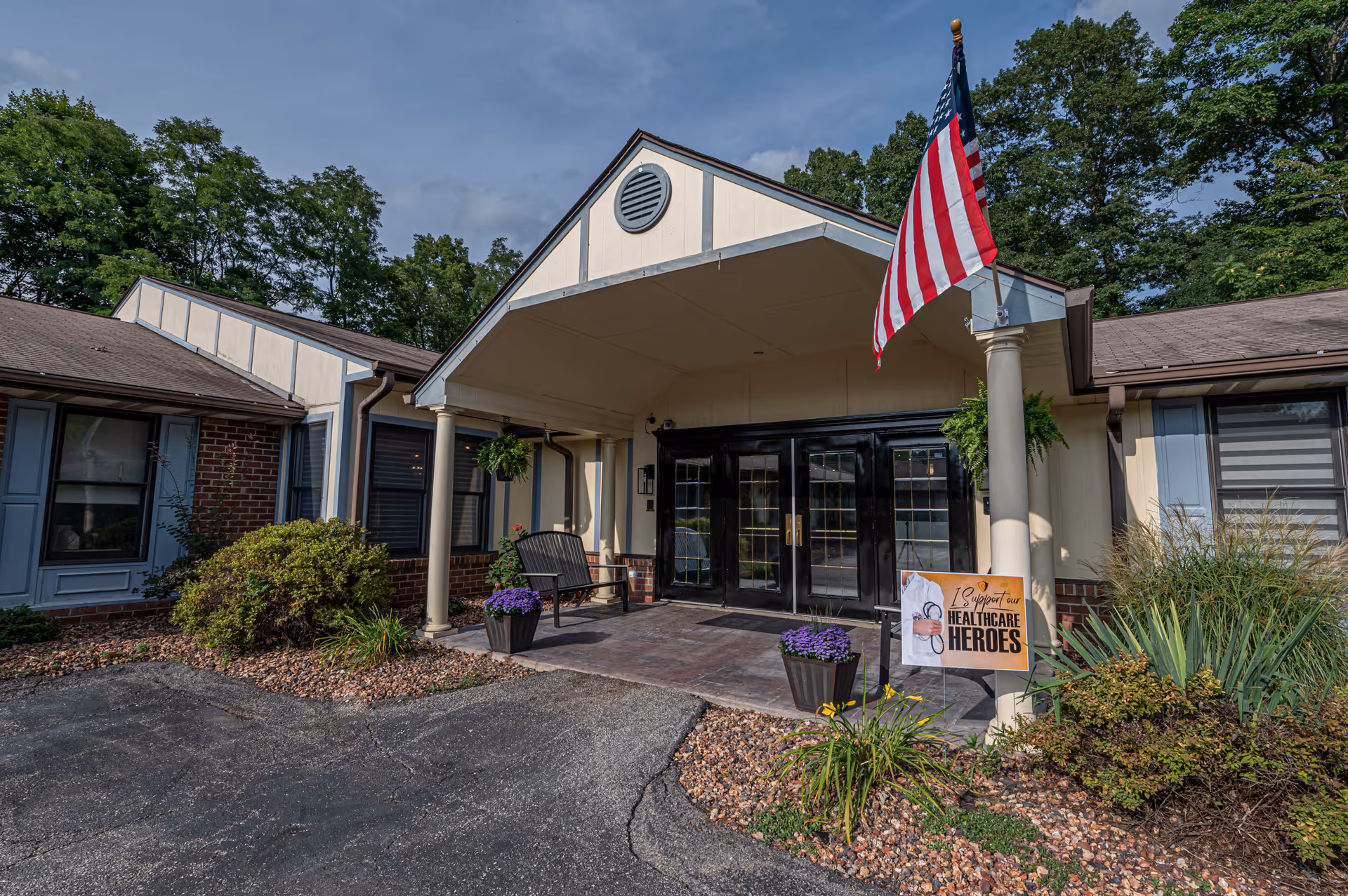 Entrance of a senior living facility with a covered porch supported by columns, an American flag, potted plants, a bench, and a sign that reads 'I Support Our Healthcare Heroes'. The building has a brick and beige exterior with blue window shutters and is surrounded by greenery and trees.