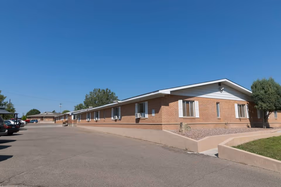 Exterior view of a single-story brick building with multiple windows and air conditioning units, a paved parking area with several parked cars, a small landscaped area with a tree, and a clear blue sky.