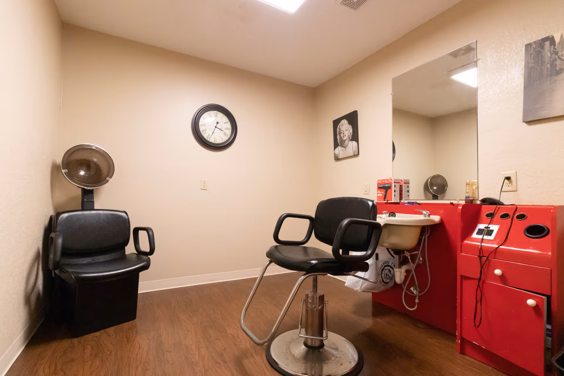 Interior of a small hair salon room with two black salon chairs, one positioned under a hair dryer and the other in front of a red counter with a sink. A large mirror is mounted above the counter, and a clock and two framed pictures hang on the beige walls. The floor is wood laminate.