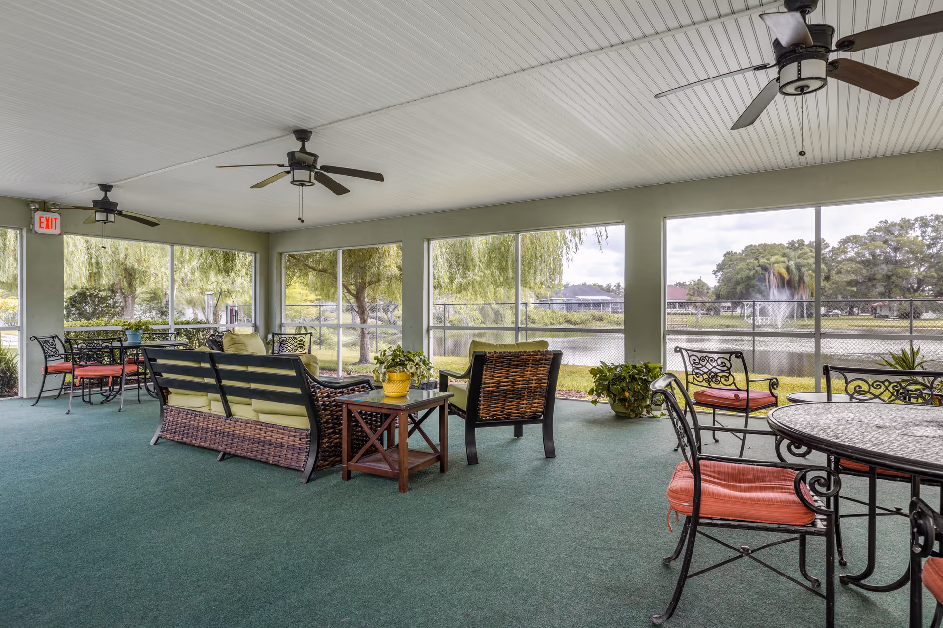 A spacious screened-in porch area with green carpet flooring, ceiling fans, and various seating arrangements including cushioned wicker sofas and metal chairs with red cushions. The porch overlooks a fenced yard with trees, a pond, and a fountain in the background.