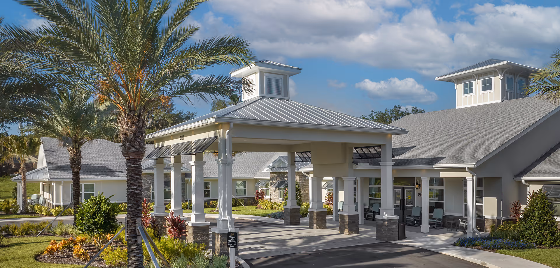Exterior view of The Addison of Narcoossee senior living facility entrance with a covered drop-off area, palm trees, and landscaped greenery under a partly cloudy sky.