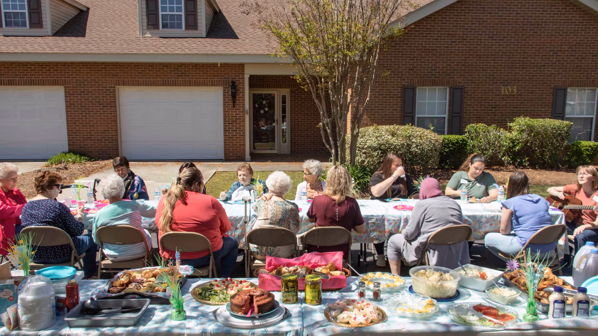 A group of elderly people and caregivers sitting around a long table outdoors in front of a brick building, enjoying a meal together. The table is covered with a floral tablecloth and filled with various dishes, including cakes, salads, and drinks. One person is playing a guitar on the right side of the image.