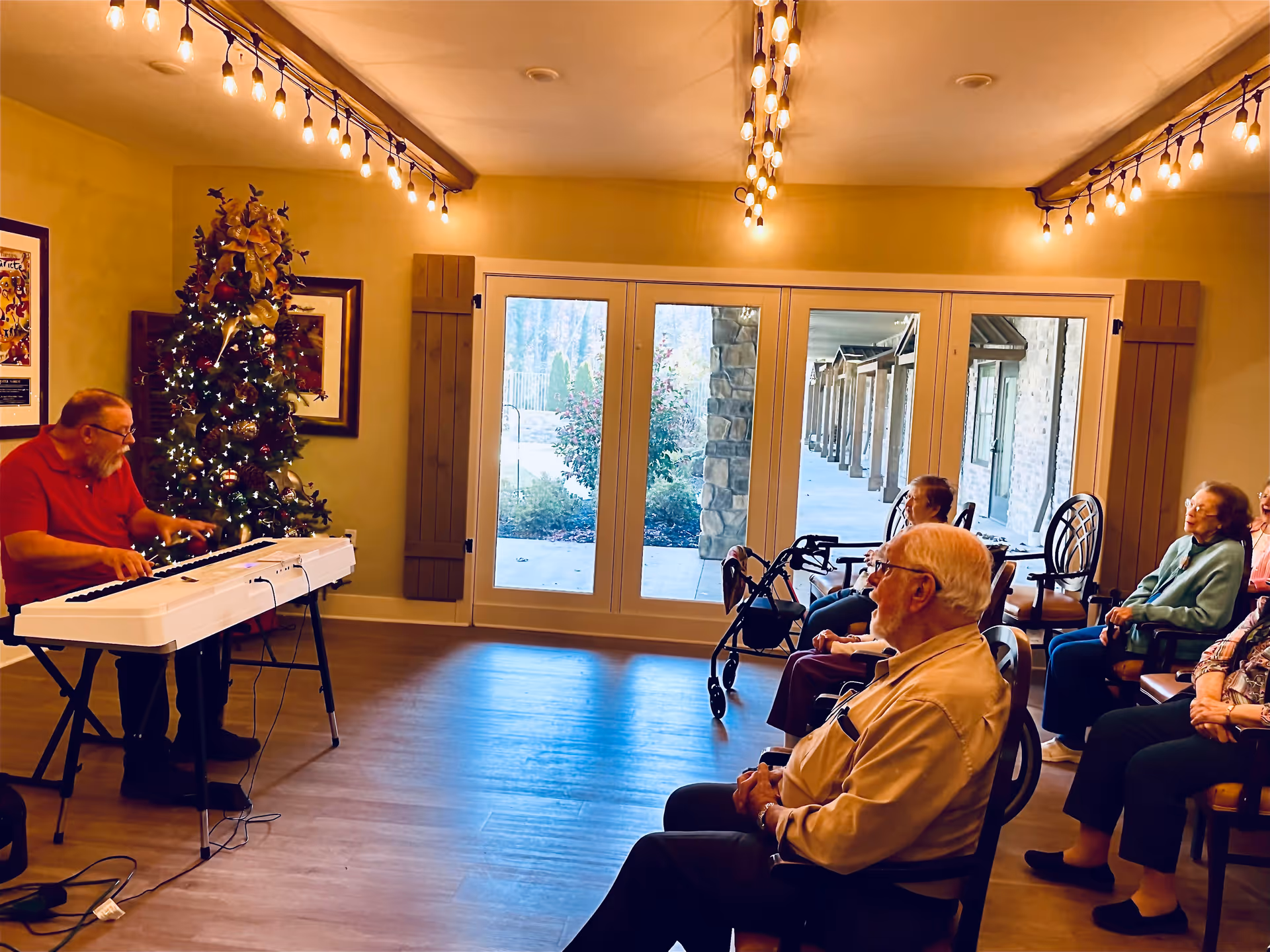 A group of elderly people seated in chairs in a room with large windows, watching a man playing a keyboard piano near a decorated Christmas tree. The room has warm lighting with string lights hanging from the ceiling.