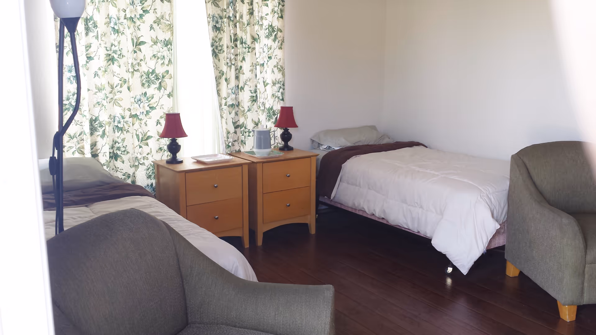 Shared bedroom with two twin beds, two wooden nightstands topped with lamps, floral curtains, and two armchairs.