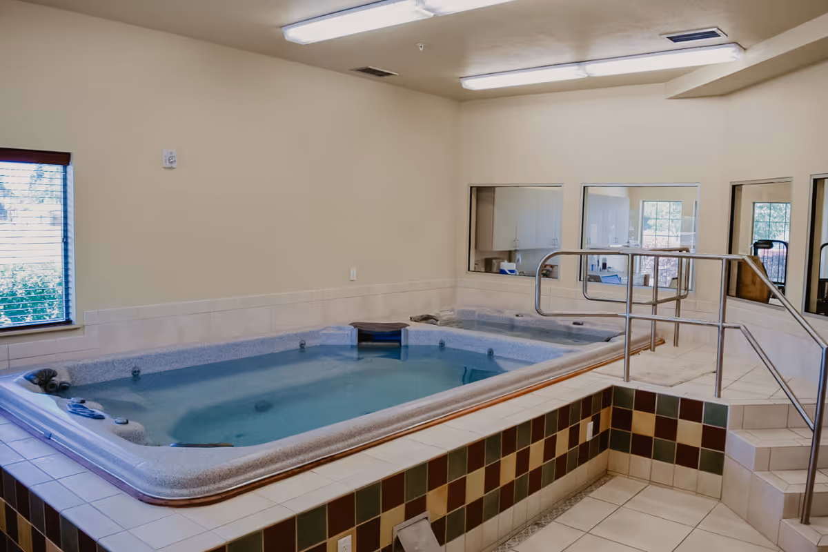 Indoor therapy pool with tiled edges and handrails, located in a room with beige walls, windows, and fluorescent ceiling lights.