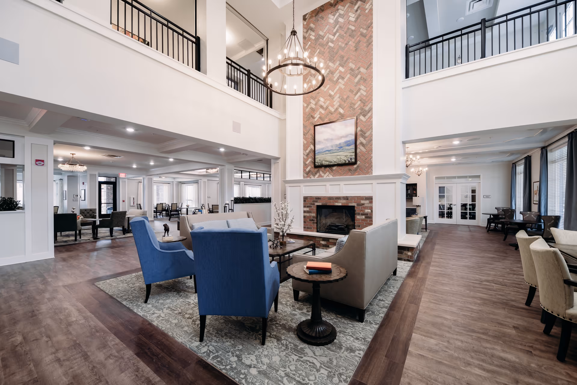 Spacious and elegant senior living facility common area with high ceilings, a large brick fireplace, and a chandelier. The room features comfortable seating including blue armchairs and beige sofas arranged around a coffee table on a patterned rug. There are additional seating areas and tables along the sides, with large windows allowing natural light to fill the space.