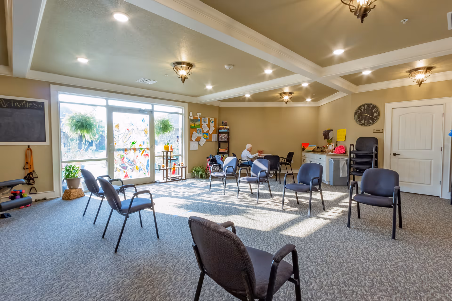 A spacious activity room in a senior living facility with several chairs arranged in a circle on carpeted floor. Large windows let in natural light, and there are plants and colorful decorations near the windows. An elderly person is seated at a table in the back corner, reading. The room has beige walls, a coffered ceiling with recessed lighting and decorative light fixtures, and a clock on the wall.
