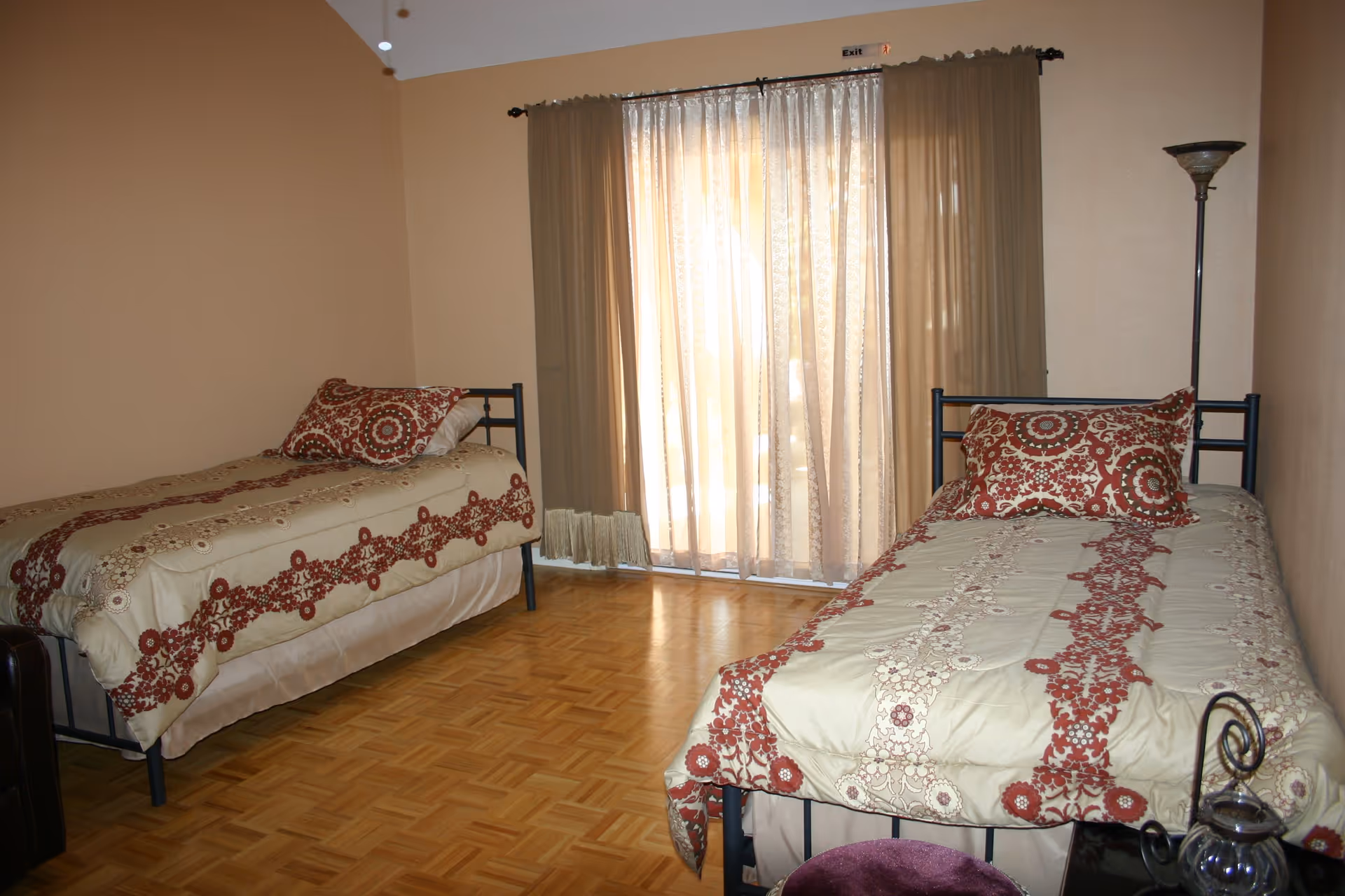 A bedroom with two single beds, each with beige and red patterned bedding and matching pillows. The room has light brown walls, a wooden parquet floor, a tall floor lamp in the corner, and a large window covered with sheer and opaque curtains allowing sunlight to filter in.