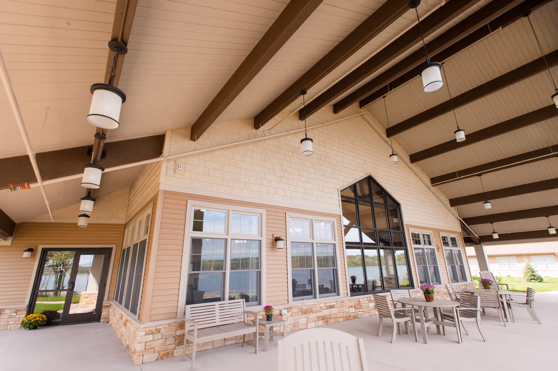 Covered outdoor patio area at Teal Lake Senior Living Community with beige siding, stone accents, multiple windows, hanging pendant lights, and several tables and chairs arranged for seating. Potted flowers decorate the tables and benches, and a glass door leads inside. A lake and greenery are visible in the background.
