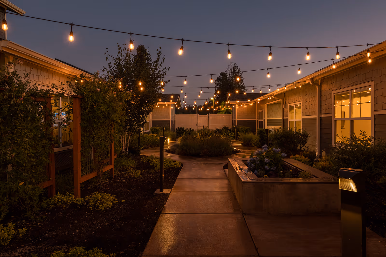 Outdoor courtyard area at dusk with string lights hanging overhead, surrounded by single-story buildings with windows. The courtyard features a paved walkway, garden beds with plants and flowers, and small trees, creating a warm and inviting atmosphere.