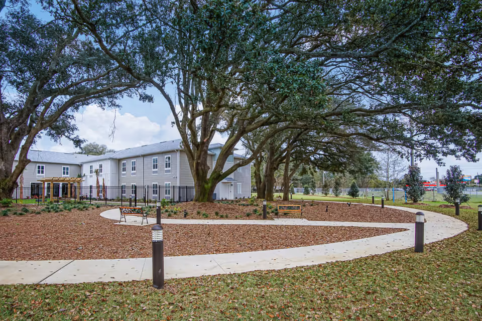 Outdoor courtyard with large oak trees, benches, a curved paved walkway, and a two-story senior living building in the background.