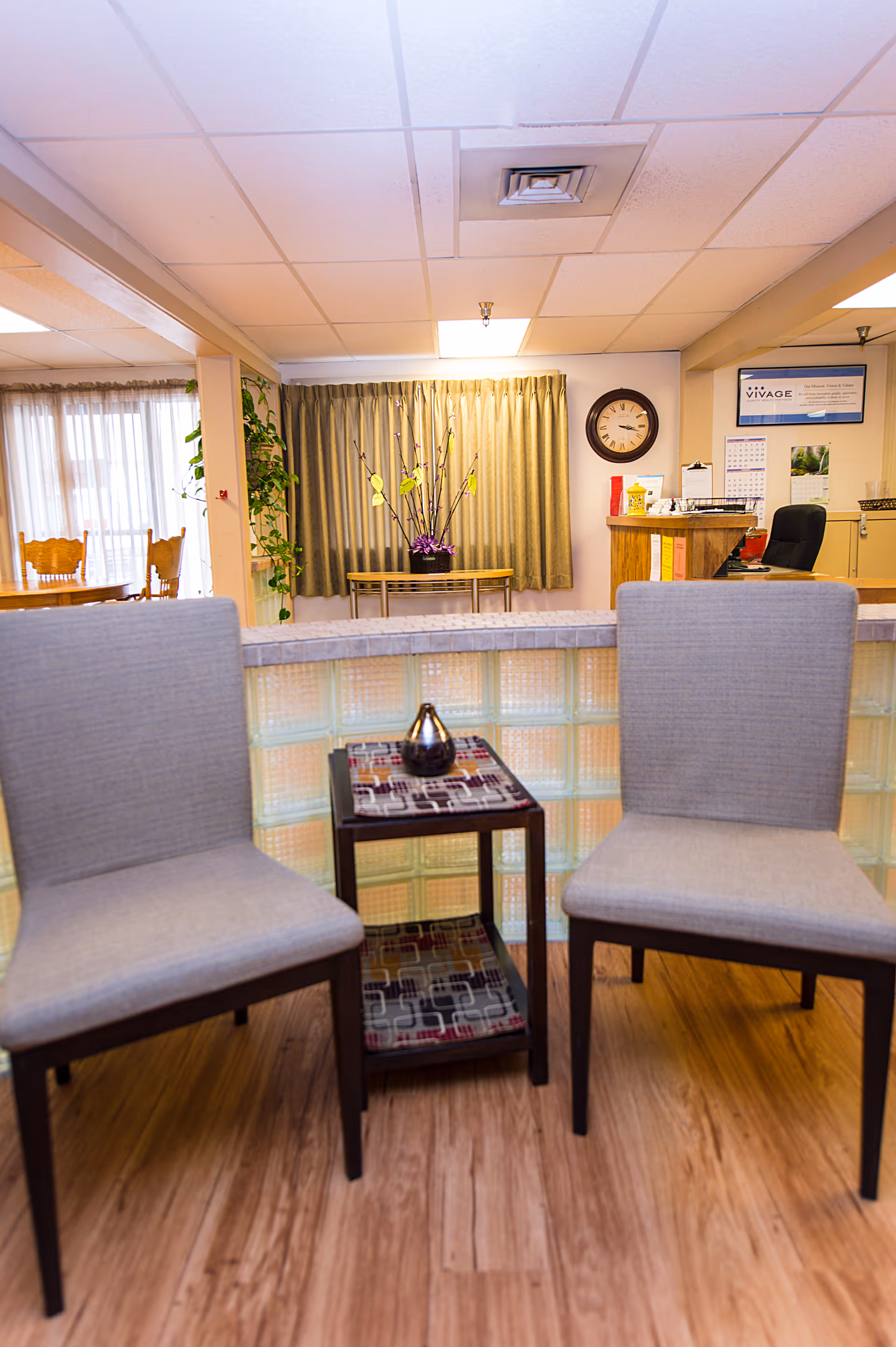 Two upholstered chairs flanking a small table in a senior living facility reception/waiting area with a clock, desk, and dining area visible in the background.