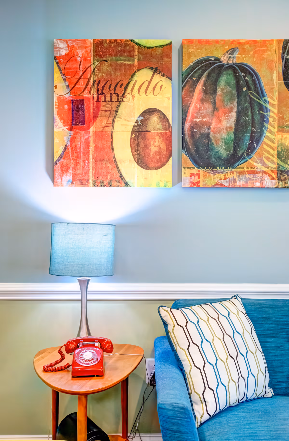 A cozy corner of a living room featuring a blue couch with a patterned pillow, a wooden side table with a retro red rotary phone, and a modern lamp with a blue shade. Above the table and couch, there are two colorful paintings depicting an avocado and a pumpkin on a light blue wall.