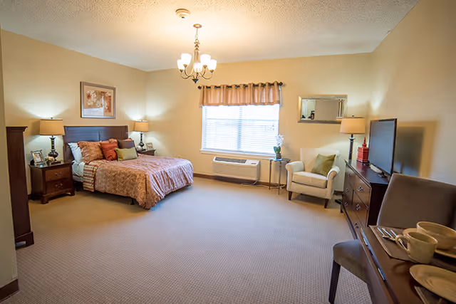 A spacious senior living bedroom with a large bed adorned with multiple pillows and a patterned bedspread. The room features two bedside tables with lamps, a window with blinds and a valance, an armchair next to a small side table with a plant, a dresser with a flat-screen TV, and a dining table with chairs and tableware. The walls are painted beige and the room is carpeted.