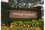 Brick sign with gold lettering reading 'Bishop Gadsden' mounted on a wall covered with green ivy, surrounded by yellow flowers and greenery.