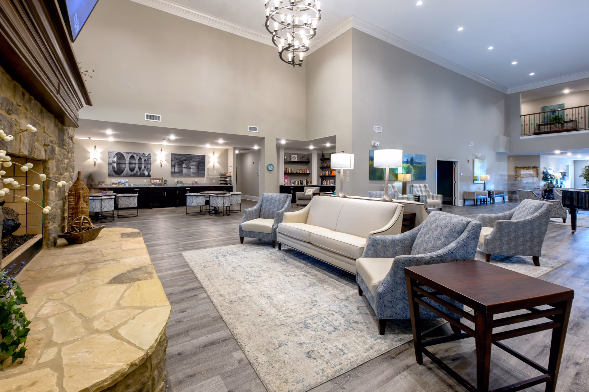 A spacious and well-lit assisted living facility common area with a high ceiling, a stone fireplace on the left, a white sofa, several patterned armchairs, a light-colored rug, wooden side tables, and modern lighting fixtures. In the background, there is a counter area with stools, artwork on the walls, and a bookshelf. The flooring is wood-style, and the overall decor is contemporary and inviting.