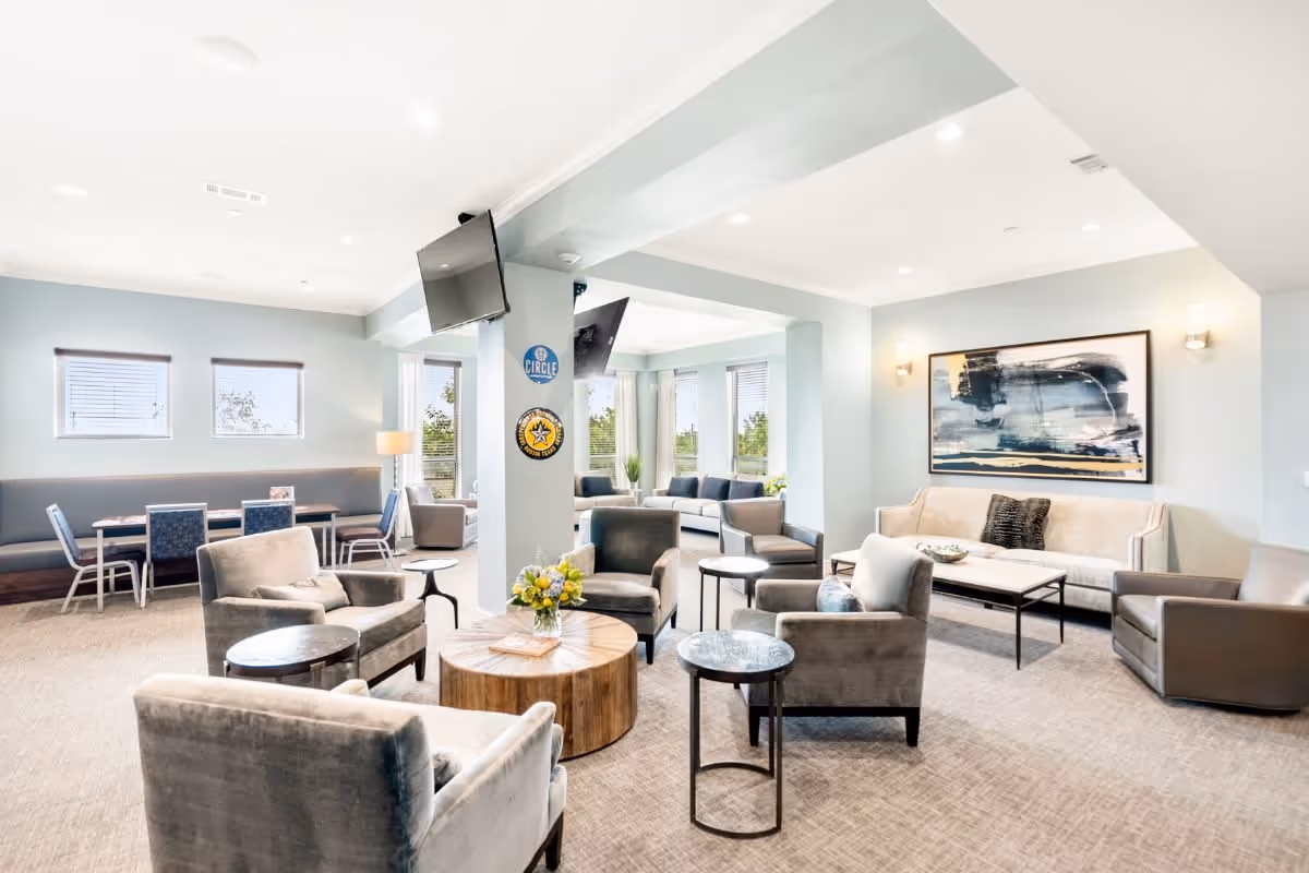 A bright and spacious living room area in a senior living facility with multiple comfortable armchairs and sofas arranged around small tables. The room features light blue walls, large windows letting in natural light, modern artwork on the wall, and two flat-screen TVs mounted on a central column. There is also a dining area with tables and chairs near the windows.