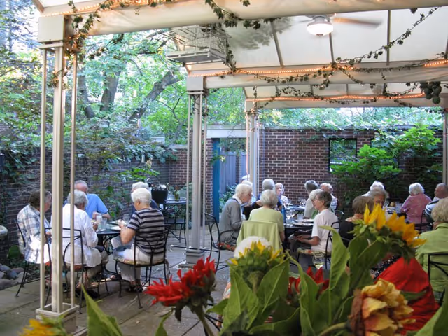 A group of elderly people sitting and socializing at tables under a covered outdoor patio area surrounded by greenery and brick walls, with flowers in the foreground.