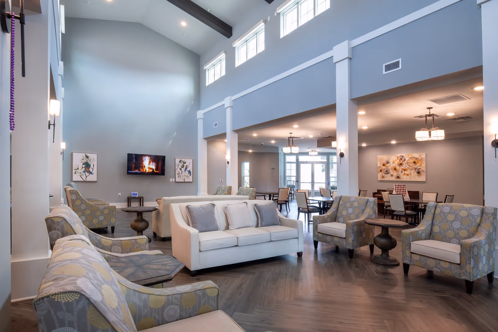 A spacious and well-lit senior living facility common area with high ceilings and multiple windows near the ceiling. The room features a white sofa with gray and white pillows, several patterned armchairs, and small wooden tables. A flat-screen TV mounted on the wall displays a fireplace scene. In the background, there is a dining area with tables and chairs, and decorative artwork on the walls.