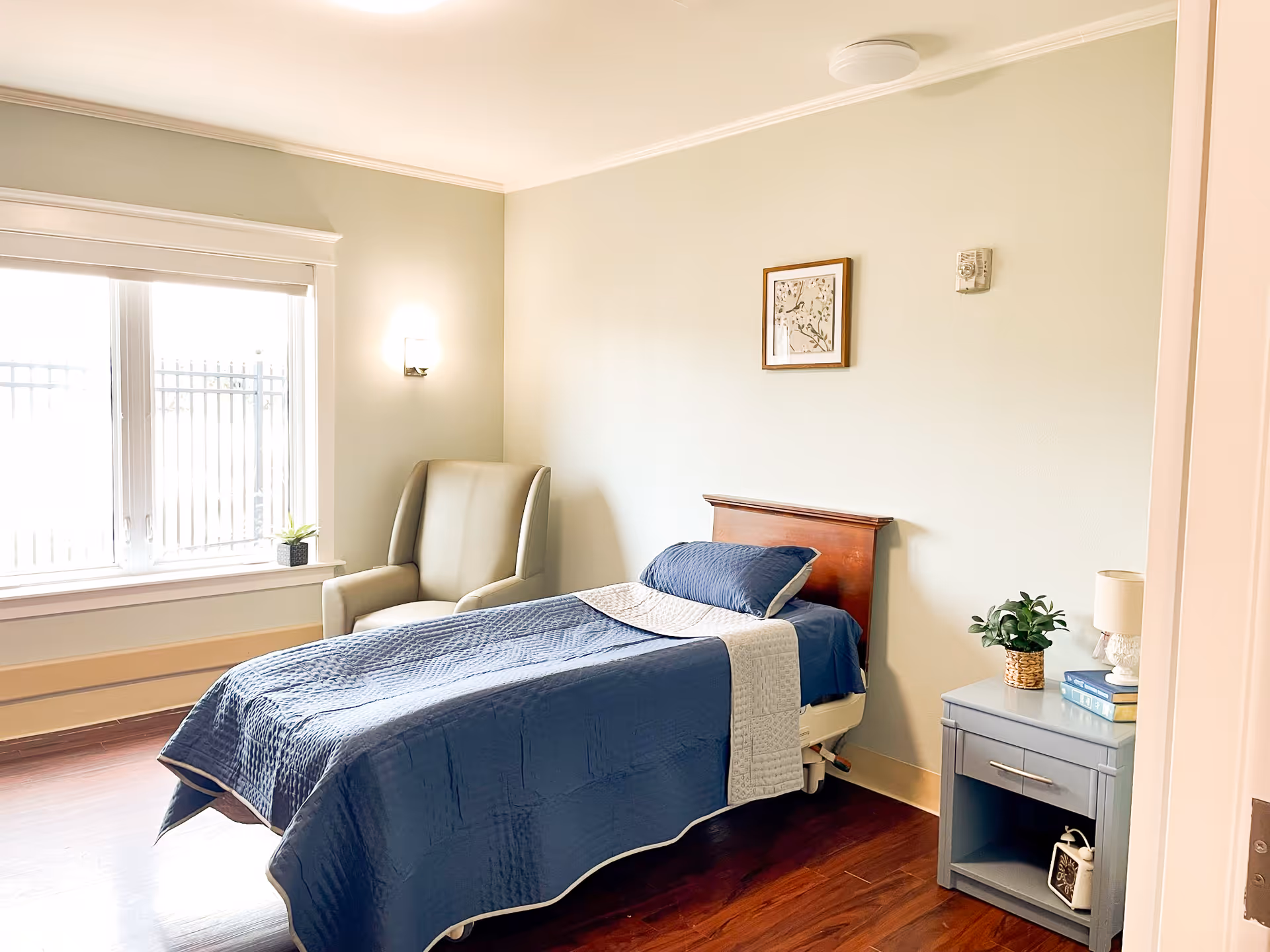 A bright and clean bedroom with a single bed covered in a blue quilt and a light gray throw. Next to the bed is a gray nightstand with a small plant, a lamp, and some books. There is a comfortable armchair near a large window with white trim, allowing natural light to fill the room. A framed picture hangs on the light-colored wall above the bed.