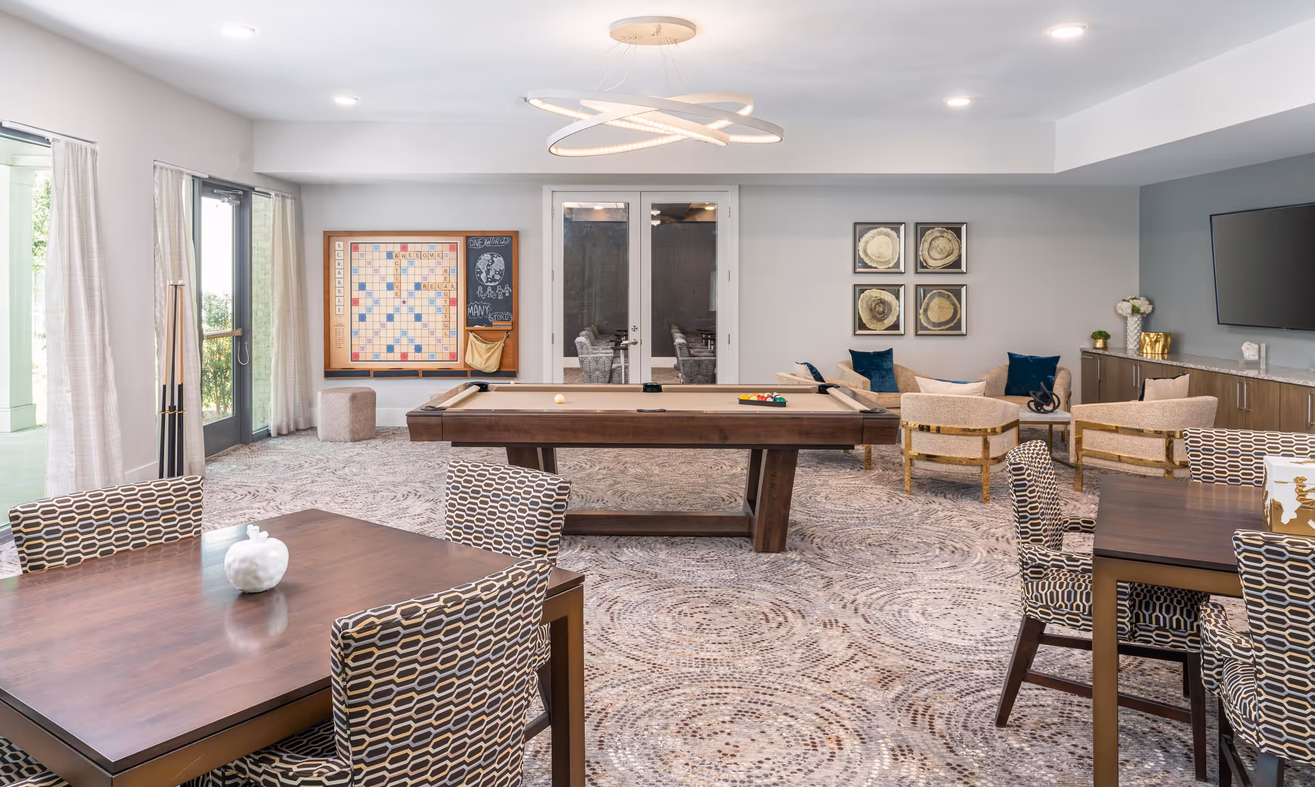 A bright and spacious game room featuring a pool table in the center, two tables with patterned chairs, a large Scrabble board on the wall, and a cozy seating area with armchairs and a sofa. The room has large windows with curtains, modern lighting, and a TV mounted on the wall above a cabinet.