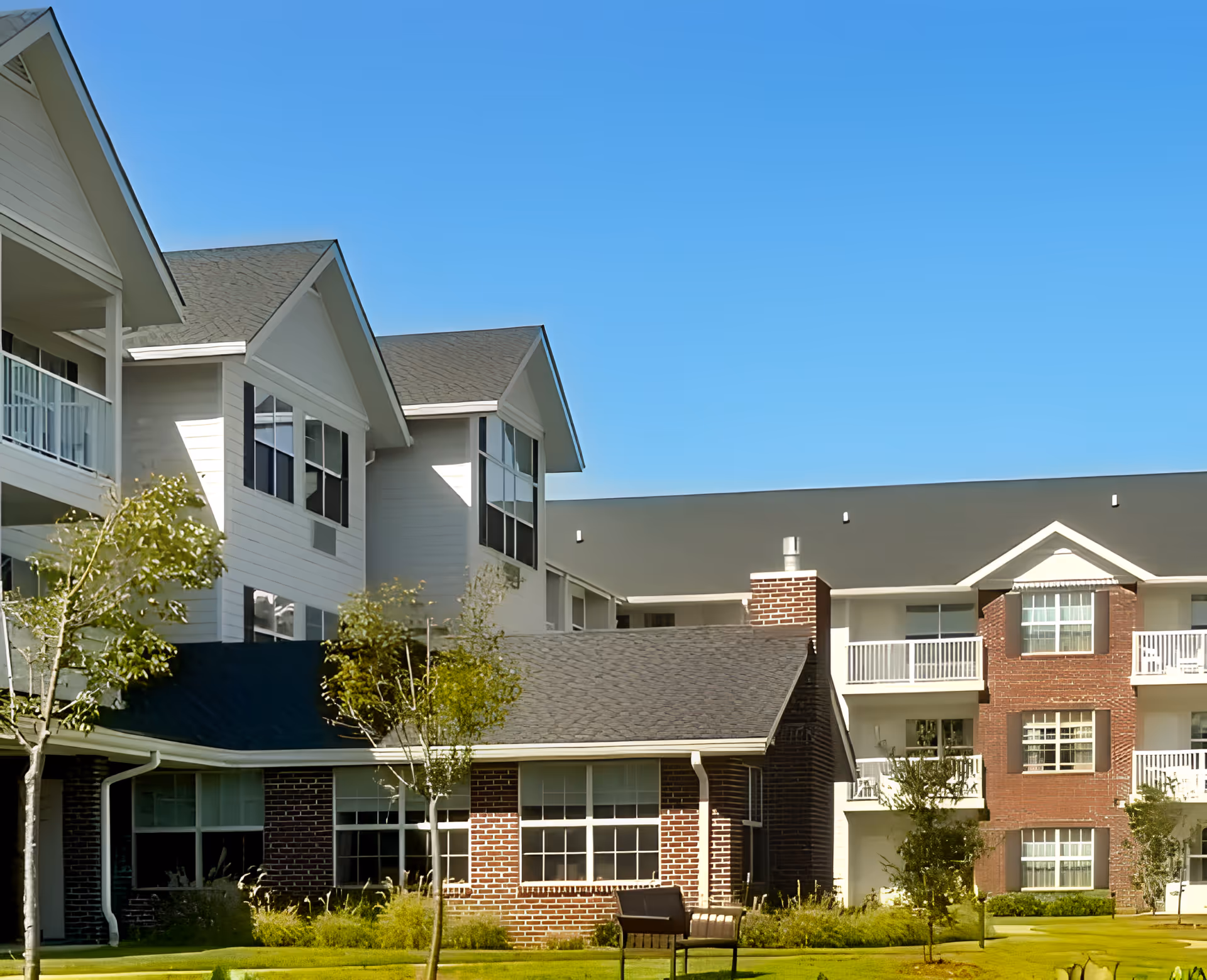 Exterior view of a senior living facility with multiple connected buildings featuring white siding and red brick facades, balconies, and large windows under a clear blue sky. There are small trees and well-maintained landscaping in the foreground.