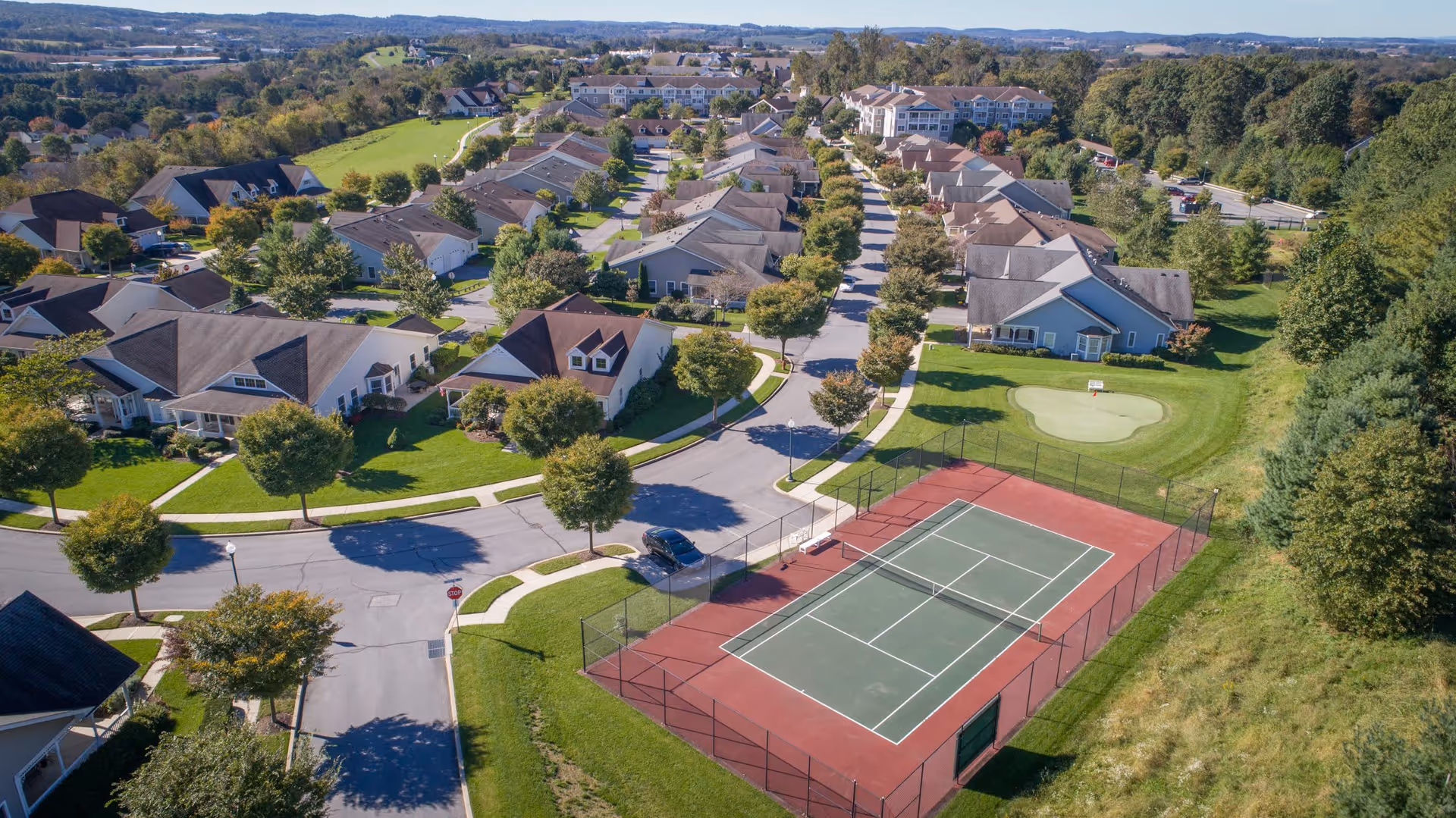 Aerial view of a residential community with rows of houses, a tennis court, and a small putting green surrounded by trees and greenery under a clear sky.