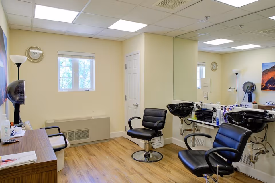 Interior of a small hair salon area with two black salon chairs in front of two black hair washing sinks. There is a large mirror on the wall behind the sinks, a wooden desk with a chair, a floor lamp, a clock on the wall, and a window letting in natural light. The room has light yellow walls and wood flooring.