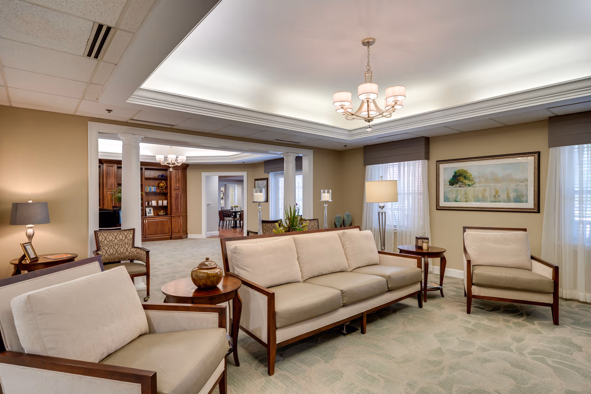 A spacious and well-lit living room area in a senior living facility with beige walls and carpet. The room features a beige sofa and two matching armchairs with wooden frames, a round wooden coffee table with a decorative item, side tables with lamps, and a framed landscape painting on the wall. In the background, there is an open area with columns, additional seating, and built-in wooden shelves with books and decor.