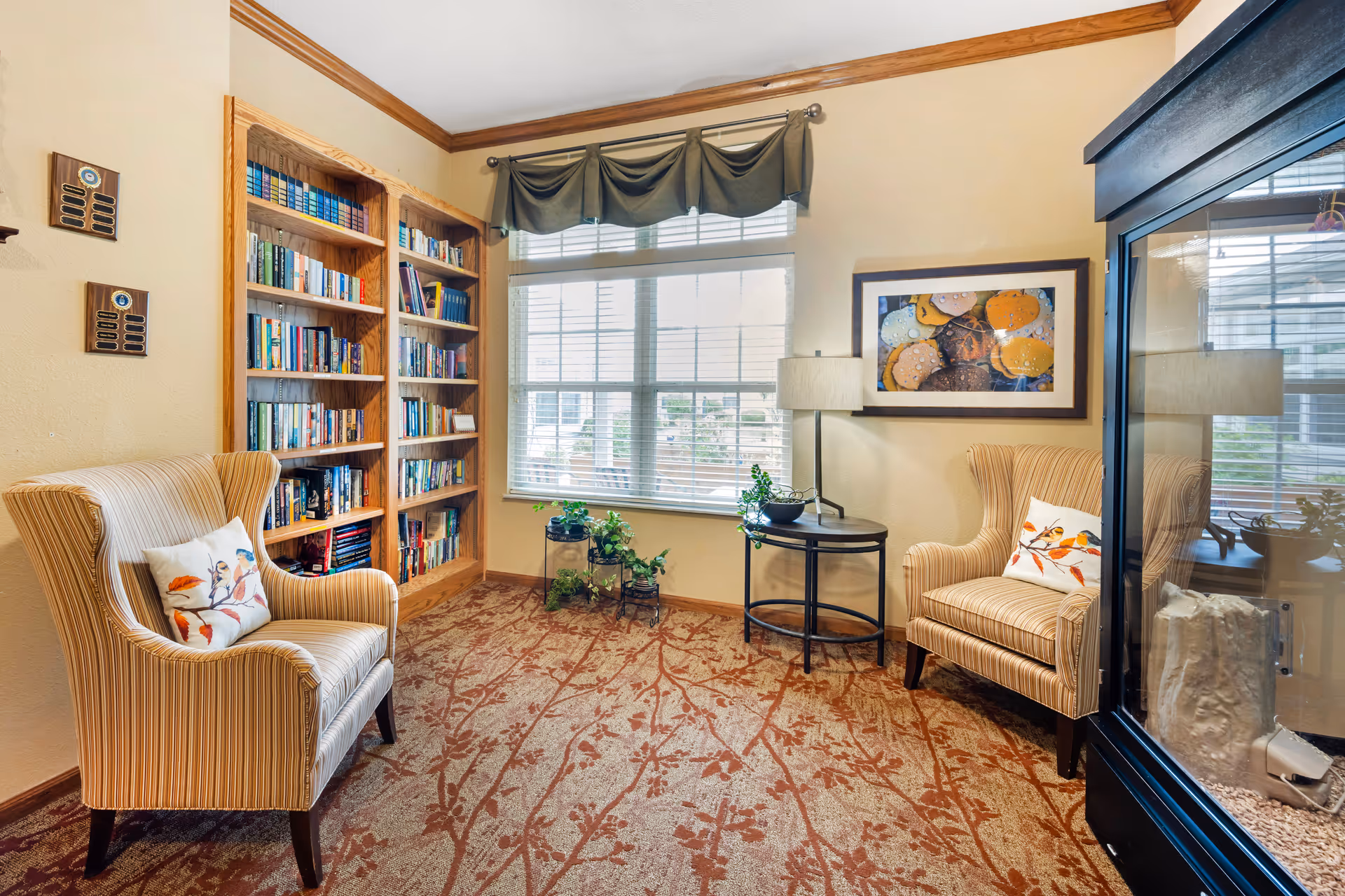 A cozy reading nook with two striped armchairs each with a decorative pillow, a wooden bookshelf filled with books, a round side table with a lamp and plants, a large window with blinds and a valance, and a framed artwork on the wall.