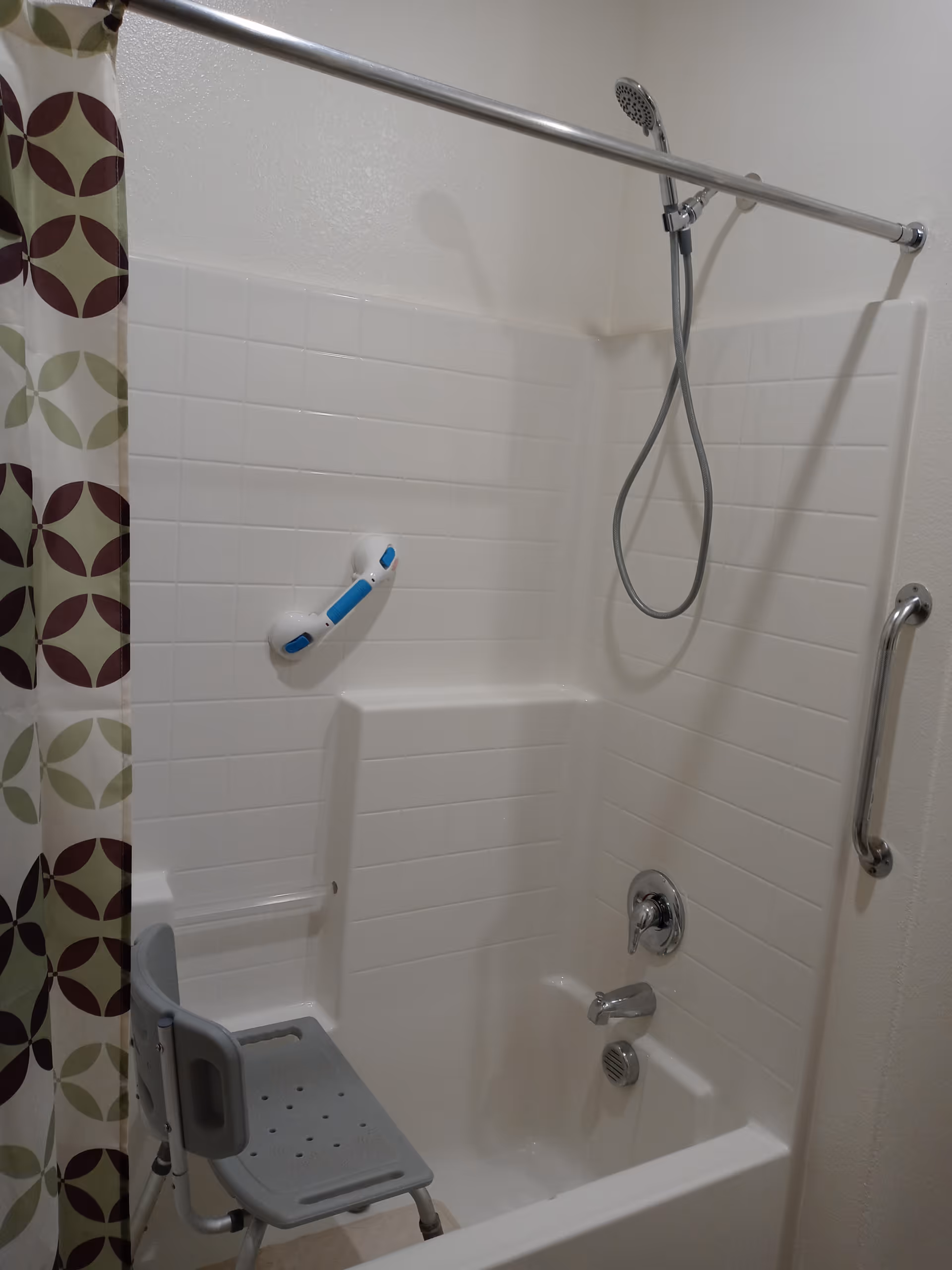 A white bathtub and shower area with a handheld showerhead, a safety grab bar on the wall, a blue and white safety handle attached to the tiled wall, and a gray shower chair inside the tub. A shower curtain with a circular pattern in green, brown, and beige colors is partially visible on the left side.